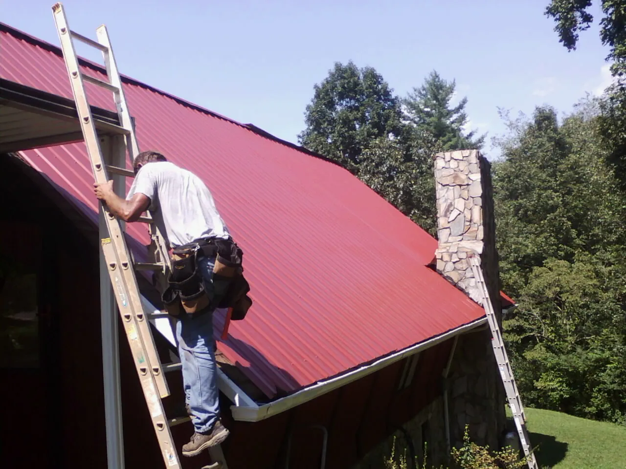 Roofer on a ladder beside a red metal roof and stone chimney — Soffit & Fascia Repair by Roofing Brothers in Adairville